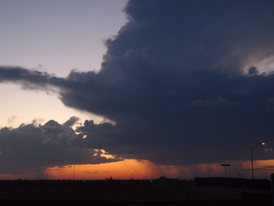 [A huge thundercloud is a dark splotch against a light-blue background. Rain streams down from the cloud to the ground. In the background of the rainstream and in sections where rain is not falling is a yellow-orange color produced by the sun setting in the distance.]